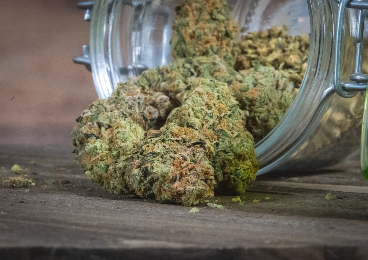 A glass jar of cannabis lying on its side with bright green buds tumbling out onto the wood table.