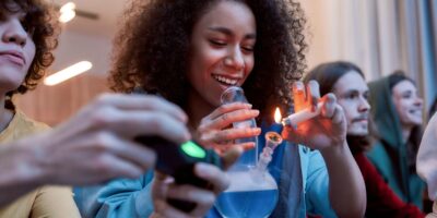 A Young Woman Lighting A Bowl Of Cannabis From A Glass Bong While Sitting On A Couch With Her Friends, Sharing Marijuana By Following The “puff Puff Pass Rule” For Proper Cannabis Etiquette.