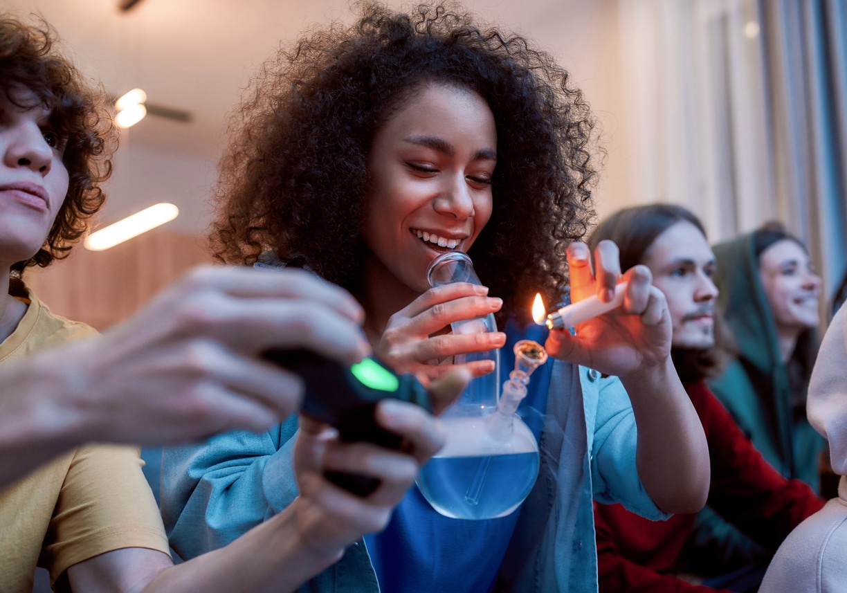 A young woman lighting a bowl of cannabis from a glass bong while sitting on a couch with her friends, sharing marijuana by following the “puff puff pass rule” for proper cannabis etiquette.